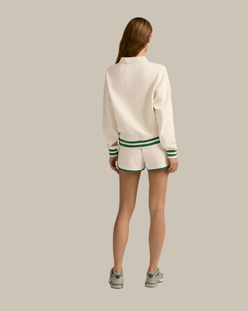 The backside view of a brunette model in front of a white background wearing a cream-colored, collared sweatshirt with a green and white striped trim and matching athletic running style shorts with green hems. The model is also wearing grey sneakers.
