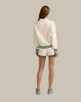 The backside view of a brunette model in front of a white background wearing a cream-colored, collared sweatshirt with a green and white striped trim and matching athletic running style shorts with green hems. The model is also wearing grey sneakers.