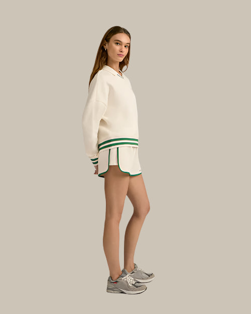 The sideview of a brunette model in front of a white background wearing a cream-colored, collared sweatshirt with a green and white striped trim and matching athletic running style shorts with green hems. The model is also wearing grey sneakers.