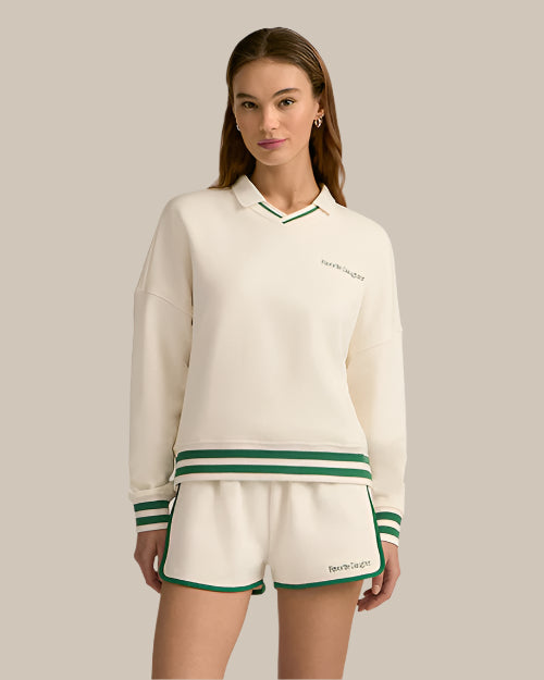 A brunette model in front of a white background wearing a cream-colored, collared sweatshirt with a green and white striped trim and matching athletic running style shorts. The sweatshirt has text on the left chest area that reads "Favorite Daughter" and the shorts have text on the left leg that also reads "Favorite Daughter".