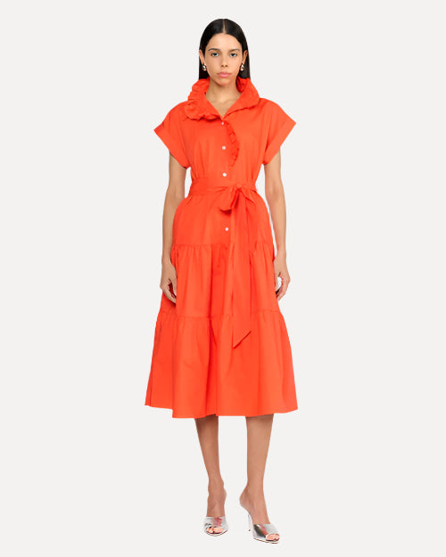 A dark-haired model wearing gold earrings and an orange dress with short, rolled sleeves, a collar, and a tied fabric belt at the waist. She is wearing silver open-toed heels in front of a white background.