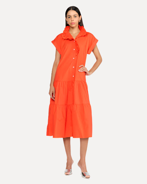 A dark-haired model wearing an orange dress with short, rolled sleeves, a collar, and white buttons down the front. She is also wearing silver open-toed heels and standing against a white background.