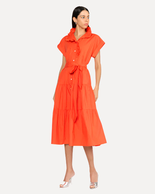 A dark-haired model wearing an orange dress with short, rolled sleeves, a collar, and a tied fabric belt at the waist. She has on silver open-toed heels and is standing in front of a white background.