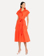 A dark-haired model wearing an orange dress with short, rolled sleeves, a collar, and a tied fabric belt at the waist. She has on silver open-toed heels and is standing in front of a white background.