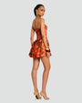 The backside view of a model wearing a ponytail, a red floral dress with thin straps, an open back and a fitted bodice, including a short, flared skirt. The model is also wearing gold hoop earrings and bangles, and gold open-toe tie up heels.
