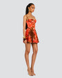 An angled view of a model standing in front of a white background wearing a red floral dress with thin straps and a fitted bodice, including a short, flared skirt. The model is also wearing gold hoop earrings and bangles, gold open-toe tie up heels and a tan bag.