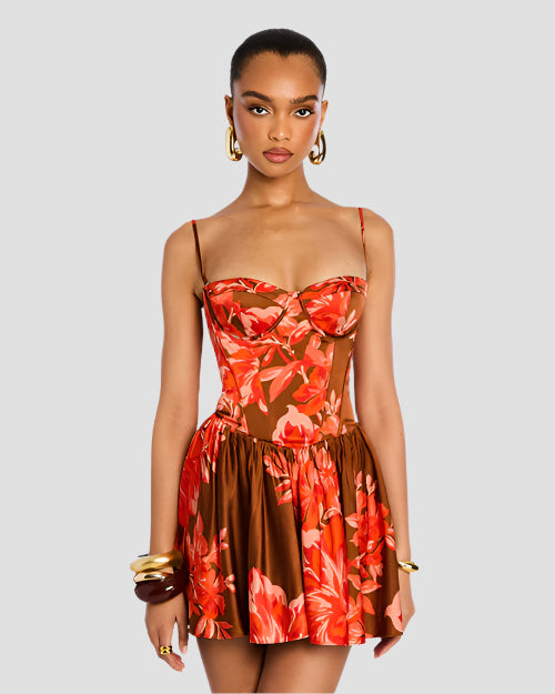 A close-up image of a model standing in front of a white background with her leg crossed over the other wearing a red floral dress with thin straps and a fitted bodice, including a short, flared skirt. The model is also wearing gold hoop earrings and bangles to match.
