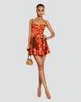 A model standing in front of a white background with her leg crossed over the other wearing a red floral dress with thin straps and a fitted bodice, including a short, flared skirt. The model is also wearing gold hoop earrings and bangles, gold open-toe tie up heels and a tan bag.