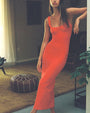 A brunette model standing in a decorative living room with plants and furniture, wearing a long form-fitting red dress with a scoop neck and thin straps.