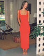 A brunette model standing barefoot inside of a decorative living room with plants and furniture. She is wearing a long form-fitting red dress with a scoop neck and thin straps.