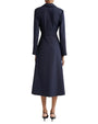 The backside view of a model wearing a long-sleeved, navy-blue dress with a tied fabric belt. The model is also wearing black pointed-toe heels with ankle straps, and standing in front of a white background.