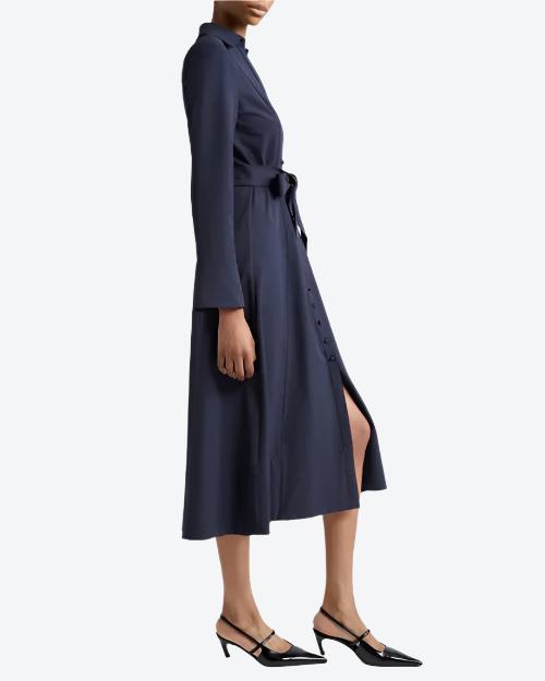 A body only sideview of a model wearing a navy long-sleeved button-down dress with a V-neck and a tied fabric bow at the waist. The model is also wearing black pointed-toe heels with ankle straps and is standing in front of a white background.
