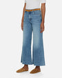 An angled lower body shot of a model wearing blue wide-leg jeans with a brown belt. The denim has a slightly faded and worn-in look and is paired with a tucked in white shirt and brown slides.