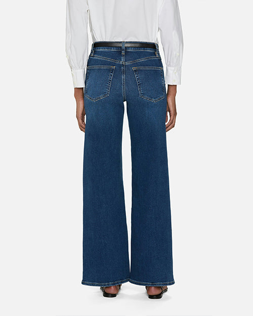 The backside view of a model standing against a plain white background wearing a white long sleeve button down. They are also wearing high-rise blue jeans with a straight-leg cut and two front pockets, featuring a medium wash. The model is wearing a black belt and black sandals.