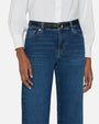A close-up waist level view of a model standing against a plain white background wearing a white long sleeve button down tucked into blue jeans with a black belt.