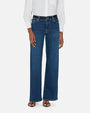 A lower body shot of a model standing against a plain white background wearing a white long sleeve button down tucked into high-rise blue jeans with a straight leg cut, a black belt and black open toe sandals.