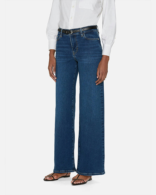 An angled lower body shot of a model standing against a plain white background, wearing a white long sleeve button down tucked into high-rise blue jeans with a straight leg cut, a black belt and black open toe sandals.