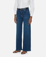 An angled lower body shot of a model standing against a plain white background, wearing a white long sleeve button down tucked into high-rise blue jeans with a straight leg cut, a black belt and black open toe sandals.