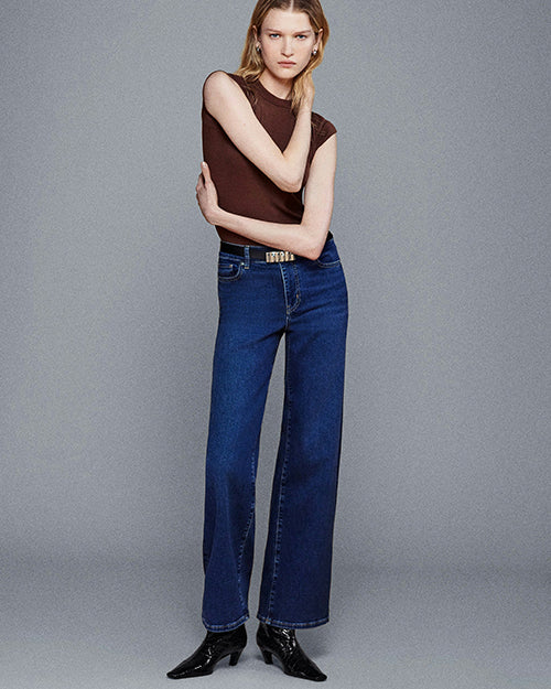 A full body shot of a model standing against a plain gray background wearing a sleeveless brown top tucked into high-rise blue jeans with a straight-leg cut and a black belt with a gold buckle, and black pointed-toe ankle boots with a low heel.