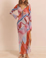 A brunette model with her hand on her hip, wearing a long-sleeve mesh dress with a vibrant abstract multicolored print, a V-neck, an open design under the chest with a bow at the center of the chest, and a side leg slid. The background is plain white.