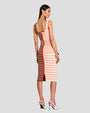 The backside view of a brunette model wearing a low-neck, below the knee-length dress with a fitted silhouette and a zipper down the back. The dress is half cream and half brown featuring horizontal stripes in shades of red and white, with two pockets and gold buttons. The model is wearing gold jewelry, white heels, and standing in front of a white background.
