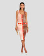 A brunette model wearing low-neck, below the knee dress, half cream and brown, featuring horizontal stripes in red and white, with red pockets and a stripe down the torso adorned by gold buttons. The model is wearing gold jewelry, a gold clutch and white high heels.