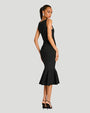 A full body backside view of a brunette model wearing a black sleeveless knee-length dress with a flared hem, including black high-heeled shoes. She is standing against a plain light background.