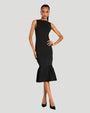 A full body view of a brunette model wearing a black sleeveless knee-length dress with a flared hem, including black high-heeled shoes. She is standing against a plain light background.