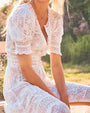 A torso-only view of a model slouching over in front of a sunny nature scene wearing a white button-down lace dress with intricate floral and delicate stripe patterns and puffed sleeves. She is also wearing a silver necklace.