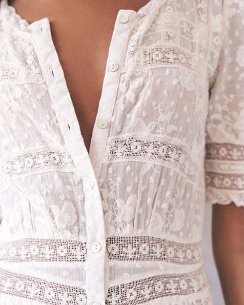 A close-up image of a models' chest area wearing a white button-down shirt with a floral lace design and a delicate horizontal lace stripe design across the body and lower sleeves.
