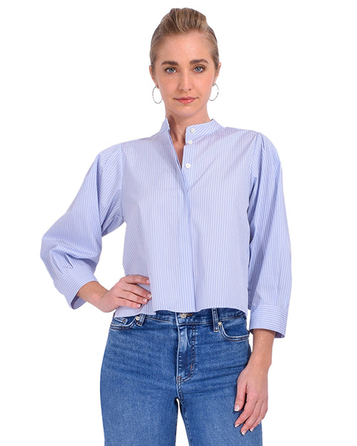 A blonde model standing against a white background is wearing a light blue and white thin striped blouse with long sleeves, a mandarin collar, and a hidden button-down design. The model is also wearing blue jeans and silver hoops.