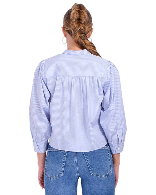A blonde model wearing a braid, standing against a white background is wearing a light blue and white thin striped blouse with long sleeves, a mandarin collar, and a hidden button-down design. The model is also wearing blue jeans and silver hoops.