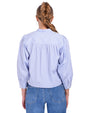 A blonde model wearing a braid, standing against a white background is wearing a light blue and white thin striped blouse with long sleeves, a mandarin collar, and a hidden button-down design. The model is also wearing blue jeans and silver hoops.
