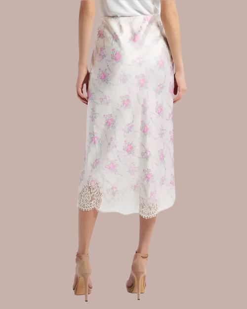 A lower-body backside view of a model wearing a long white skirt with delicate floral patterns and lace detailing at the hem. The person is also wearing beige high-heeled shoes and standing in front of a grey background.