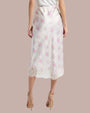 A lower-body backside view of a model wearing a long white skirt with delicate floral patterns and lace detailing at the hem. The person is also wearing beige high-heeled shoes and standing in front of a grey background.