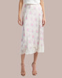A lower body frontside view of a model wearing a long white skirt with delicate floral patterns and lace detailing at the hem. The person is also wearing beige open-toed high-heeled shoes and standing in front of a grey background.
