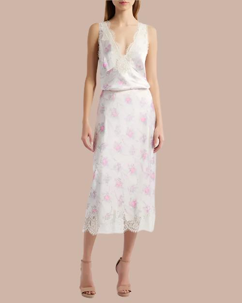 A full body view of a model wearing a white floral-patterned tank top adorned with pink flowers, a V-neck, and delicate lace trim with scalloped edges along the neckline and straps. The model is wearing long matching skirt, and brown heels while standing against a beige background.