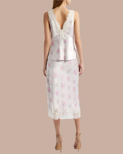 A full body backside view of a model wearing a white floral-patterned tank top adorned with pink flowers, a V-back, and delicate lace trim with scalloped edges along the neckline and straps. The model is wearing long matching skirt, and brown heels while standing against a beige background.