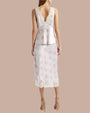 A full body backside view of a model wearing a white floral-patterned tank top adorned with pink flowers, a V-back, and delicate lace trim with scalloped edges along the neckline and straps. The model is wearing long matching skirt, and brown heels while standing against a beige background.
