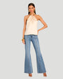 A brunette model standing in front of a white background, wearing a beige strapless halter top with a pleated design at the neckline, connected to a colorful oval beaded necklace as part of its design. The top is paired with high-waisted blue jeans, high heels, and a chunky gold bracelet and earrings.