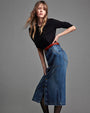 A sideview of a brunette model in a  bent over pose, standing against a plain grey background and wearing a black long-sleeve top tucked into a shin-length blue denim skirt with gold buttons down the front, and black tights under.