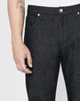 A close-up waist level view of a model with a dolphin tattoo on his forearm wearing black jeans with silver buttons tucked into a white shirt. The background is plain white.
