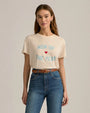 A torso level shot of a brunette model wearing a beige crewneck t-shirt with the text "MOM OF THE YEAR" written in blue, and a small red heart. The model is also wearing high-waisted blue jeans and a brown belt, against a grey background.
