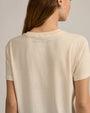 A close-up backside view of a brunette model with her hair swept to the side, wearing silver hoop earrings and a beige crewneck t-shirt with the words "Favorite Daughter" in light blue below the neckline.