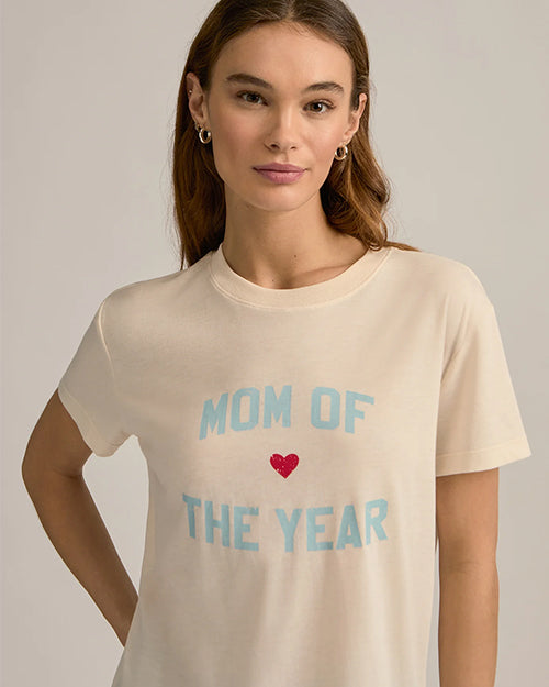 A brunette model wearing silver hoop earrings and a beige crewneck t-shirt with the text "MOM OF THE YEAR" written in in blue, and a small red heart.