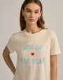 A brunette model wearing silver hoop earrings and a beige crewneck t-shirt with the text "MOM OF THE YEAR" written in in blue, and a small red heart.