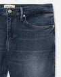 A close-up hip-level view of a pair of blue jeans with a silver button and zipper closure, and a white rectangular tag with the word "FRAME" printed on it. The background is plain white.