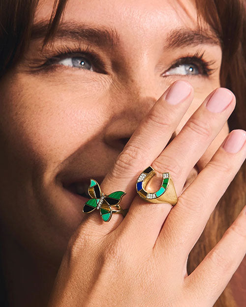 A zoomed in image of a models' laughing face covered by her hand, wearing a gold ring with a horseshoe design, showcasing divided multi-color gems and a gold outline, and a butterfly shaped ring with the same design.