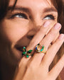 A zoomed in image of a models' laughing face covered by her hand, wearing a gold ring with a horseshoe design, showcasing divided multi-color gems and a gold outline, and a butterfly shaped ring with the same design.