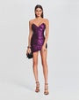 A full body view of a brunette model wearing a strapless, shiny purple dress with ruched detailing all across it and a sweetheart neckline. The model is also holding a black clutch and wearing open-toed black heels to match and is standing in front of a white background.